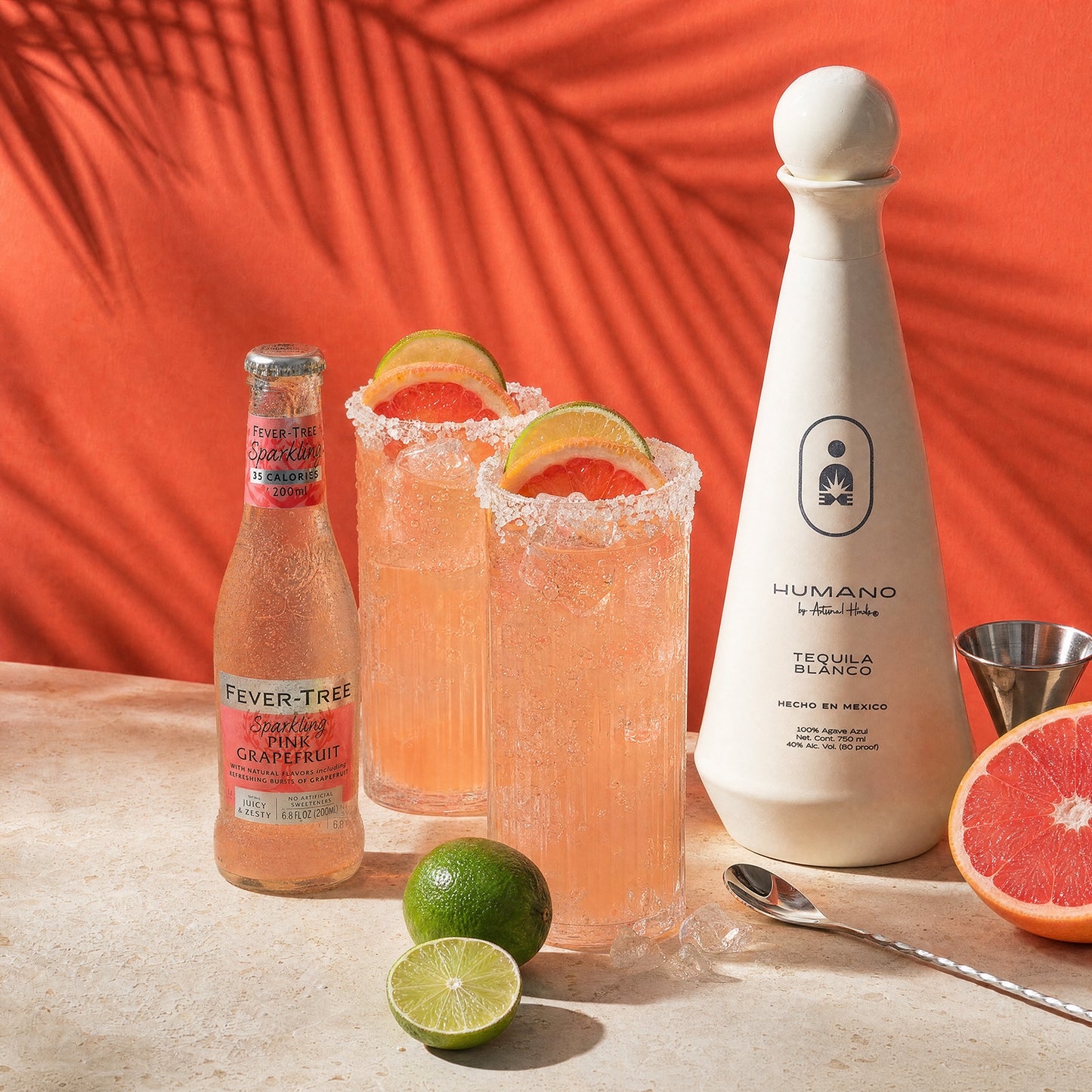 A styled product shot featuring a bottle of Humano tequila blanco and a bottle of Fever Tree sparkling citrus cocktail, alongside two pink cocktails with sugar-rimmed glasses and lime and grapefruit garnishes on a stone surface against a bright orange backdrop with palm leaf shadows.