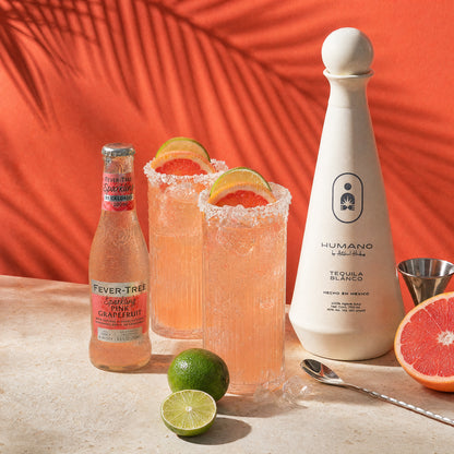 A styled product shot featuring a bottle of Humano tequila blanco and a bottle of Fever Tree sparkling citrus cocktail, alongside two pink cocktails with sugar-rimmed glasses and lime and grapefruit garnishes on a stone surface against a bright orange backdrop with palm leaf shadows.