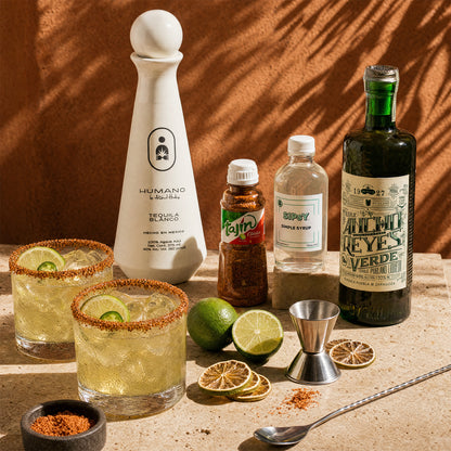 A styled bar scene with two filled margarita glasses rimmed with spice and lime, a white tequila bottle labeled Humano Tequila Blanco, a bottle of Tajín seasoning, a bottle of simple syrup, a dark green bottle of Ancho Reyes Verde, a metal jigger, a spoon, and lime/lemon slices on a stone surface against a warm textured backdrop.