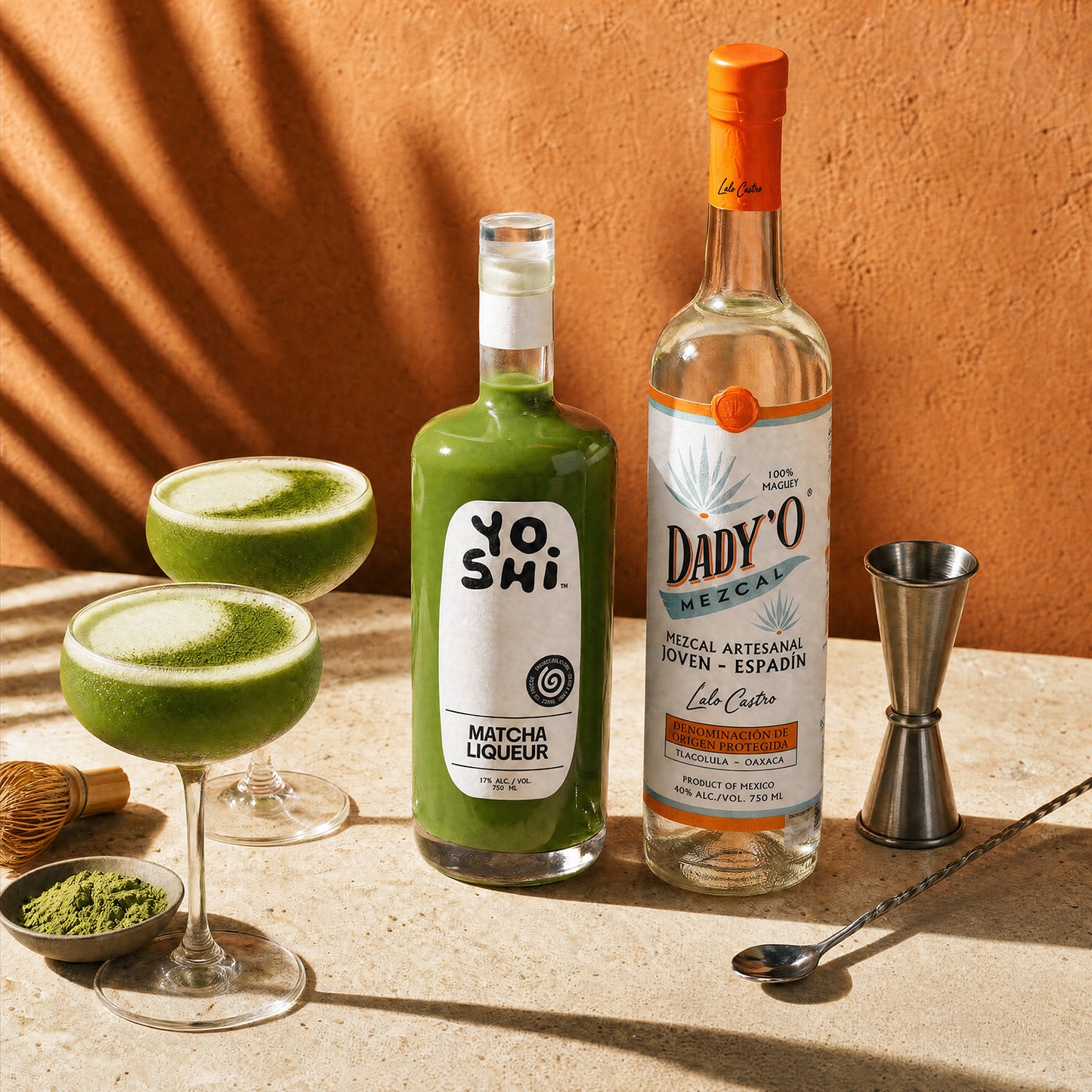 A styled product shot featuring a bottle of Yoshi Matcha Liqueur and a bottle of Dady'O Mezcal, alongside two green cocktails in coupe glasses, a metal jigger, a spoon, and small bowls of matcha powder, set against a warm terracotta wall with leaf shadows.