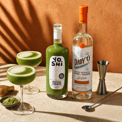 A styled product shot featuring a bottle of Yoshi Matcha Liqueur and a bottle of Dady'O Mezcal, alongside two green cocktails in coupe glasses, a metal jigger, a spoon, and small bowls of matcha powder, set against a warm terracotta wall with leaf shadows.