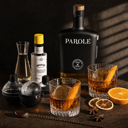 A Parole brand black bottle of rum or whiskey stands on a dark wooden table beside two rocks glasses filled with amber liquor, orange slices, star anise, a decanter, ice balls, and a bar spoon. The scene is styled with dramatic warm lighting and shadows against a dark background.