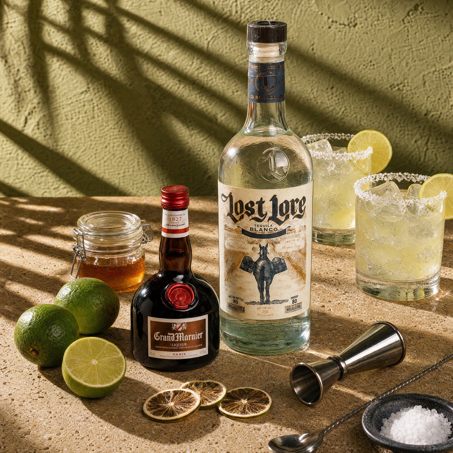 A styled bar scene with a bottle of Lost Lore Blanco tequila, a bottle of Grand Marnier cognac, two prepared cocktails with lime wheels, limes, dried lime slices, a jigger, and a small bowl of salt on a textured surface against a sunlit wall with leaf shadows.