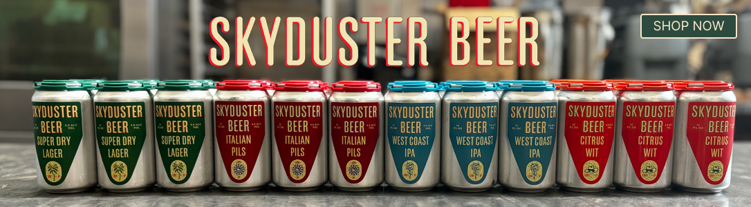 A row of colorful Skyduster Beer cans lined up on a counter with a blurred bar or brewery background. Large text at the top reads 'SKYDUSTER BEER' and a green 'SHOP NOW' button appears in the top right.